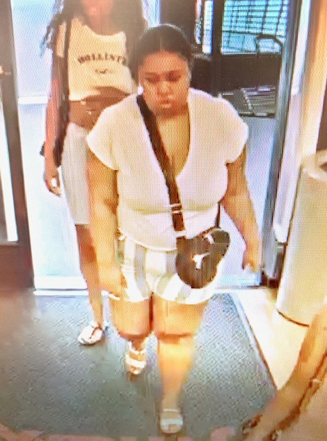 Photo of second female suspect wearing light color v-neck t-shirt with gray and white striped shorts, white sandals and a black cross-body purse. 